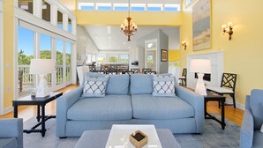 A spacious living room with a large sofa, a coffee table, and a fireplace. There is a dining area in the background with a large window. - THIS FABULOUS FIRST-TIER PLANTATION property is the ultimate family gathering place! (St. George Island)