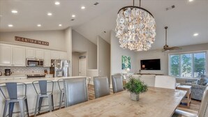 Uma cozinha moderna com armários brancos, uma ilha grande com banquetas, um lustre e um espaço de jantar com uma mesa ampla.