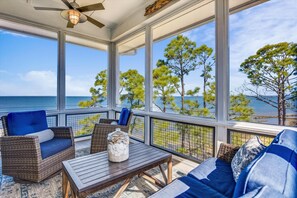 A covered outdoor seating area with wicker furniture, a table, and a view of the ocean and trees.