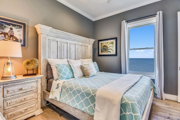 A bedroom with a large bed, a dresser, a lamp, and a view of the sea through a window.