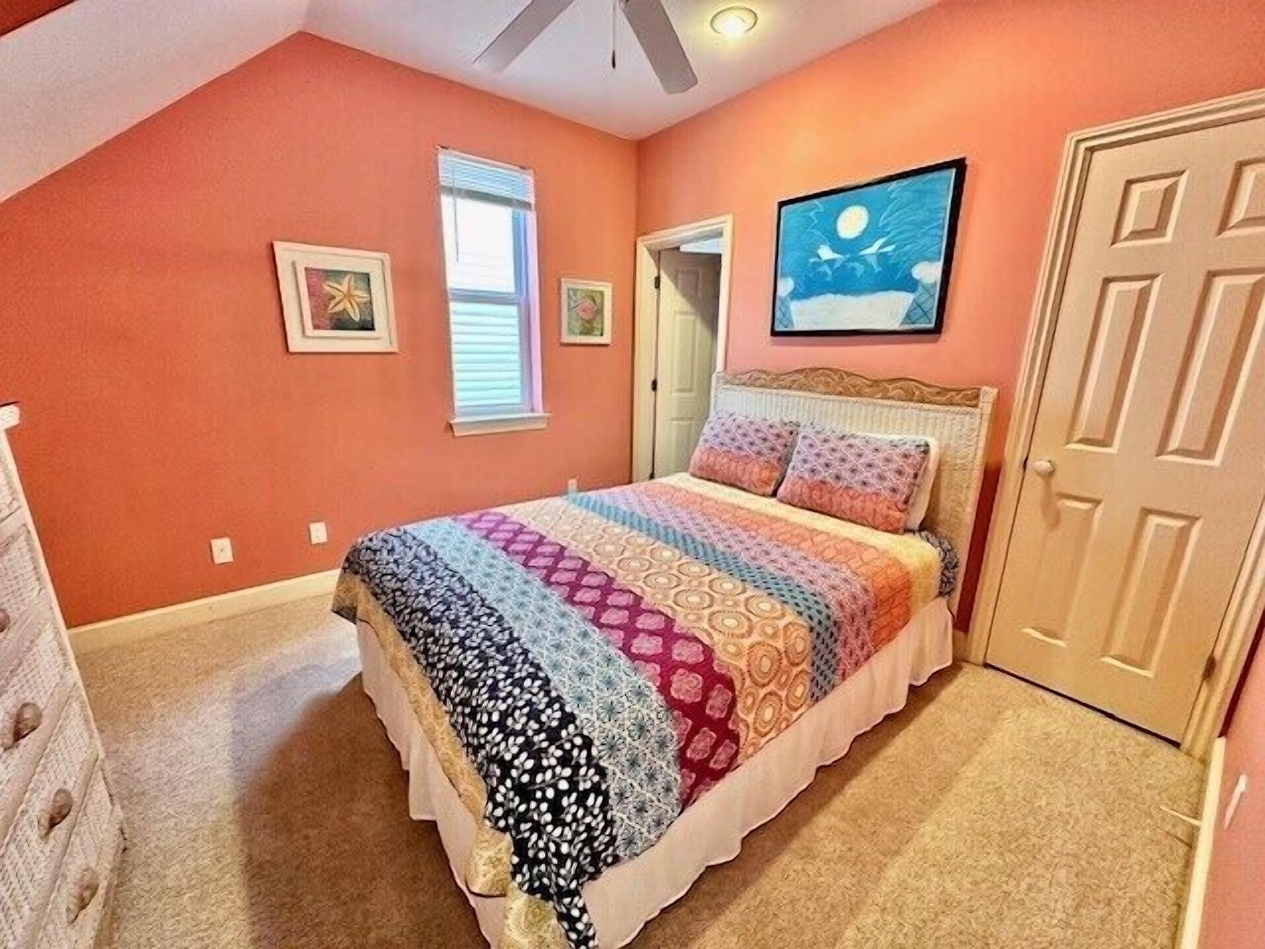 Een slaapkamer met een schuine plafond, een bed met een kleurrijke quilt, twee ingelijste foto’s aan de muur en een deur aan de rechterkant.