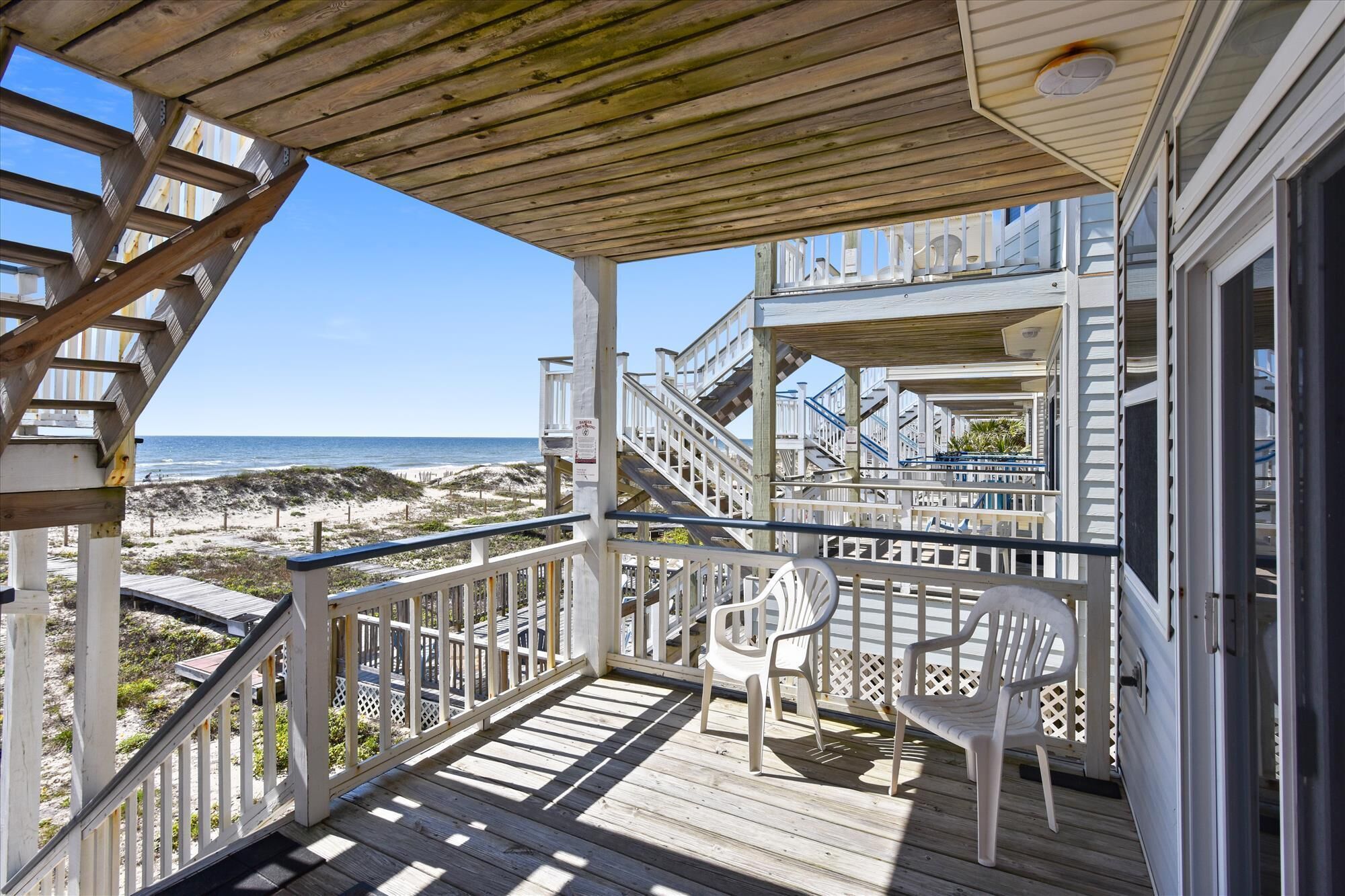A wooden deck with a view of the beach and ocean. There are stairs leading to a balcony with a railing.