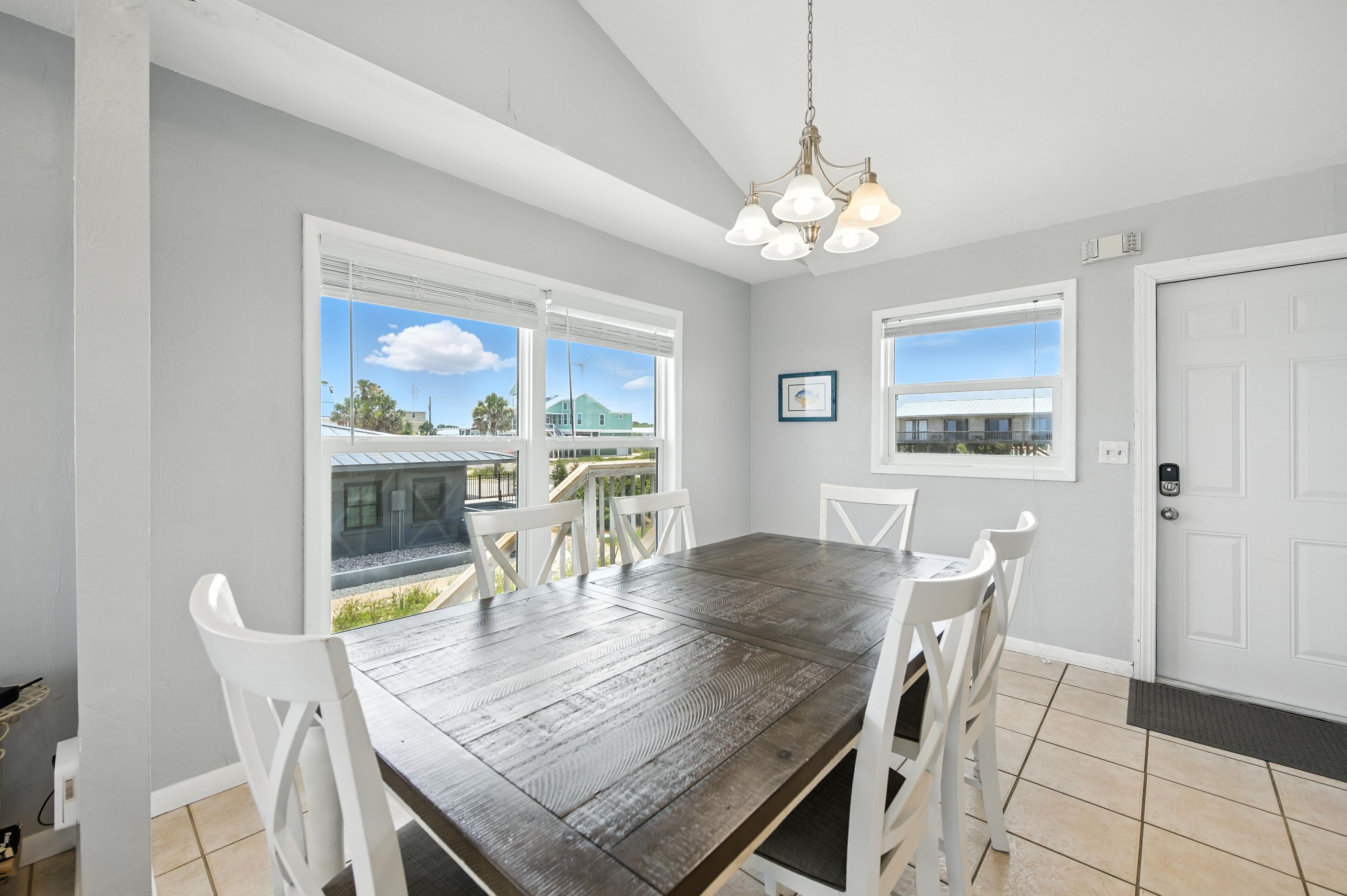 A dining area with a wooden table and white chairs, large windows with a view of a house and trees, and a white door with a keypad lock.