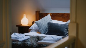 Ein ordentlich bezogenes Bett mit einem gemusterten, einem weißen und einem dunklen Kopfkissen. Ein Holz-Kopfteil und ein Nachttisch mit einer weißen Vase und einer Lampe sind zu sehen.