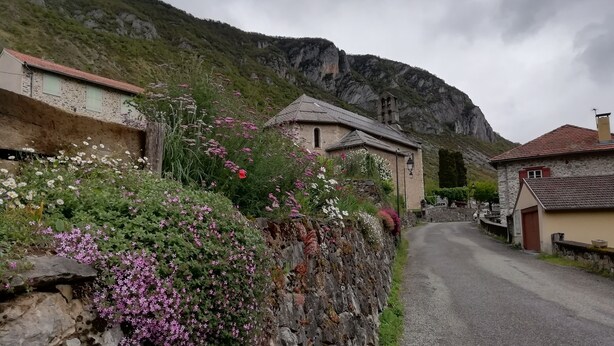 A narrow road flanked by stone walls and buildings with stone facades and red-tiled roofs.