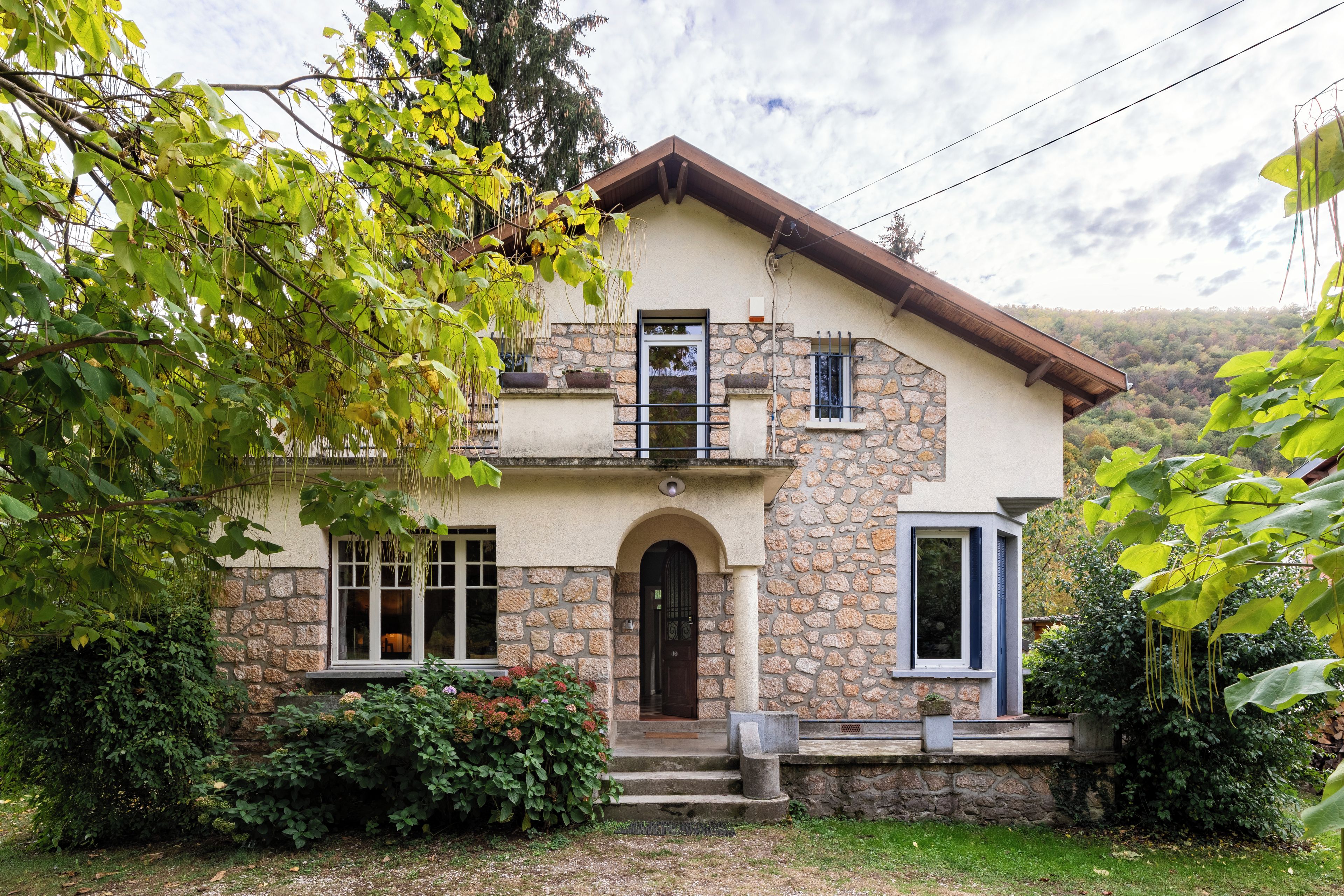 A two-story stone house with a balcony, a front door, and a small garden area.