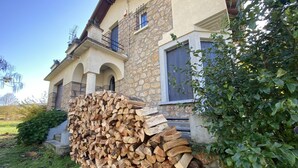 A house with a stone facade, a wooden log pile, and a green bush.