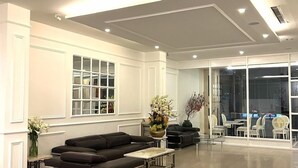 A spacious living area with a large sectional sofa, a coffee table, and a vase of flowers. There are multiple windows and a view of a dining area with tables and chairs. - Dyn Opera Hotel (Ho Chi Minh City)