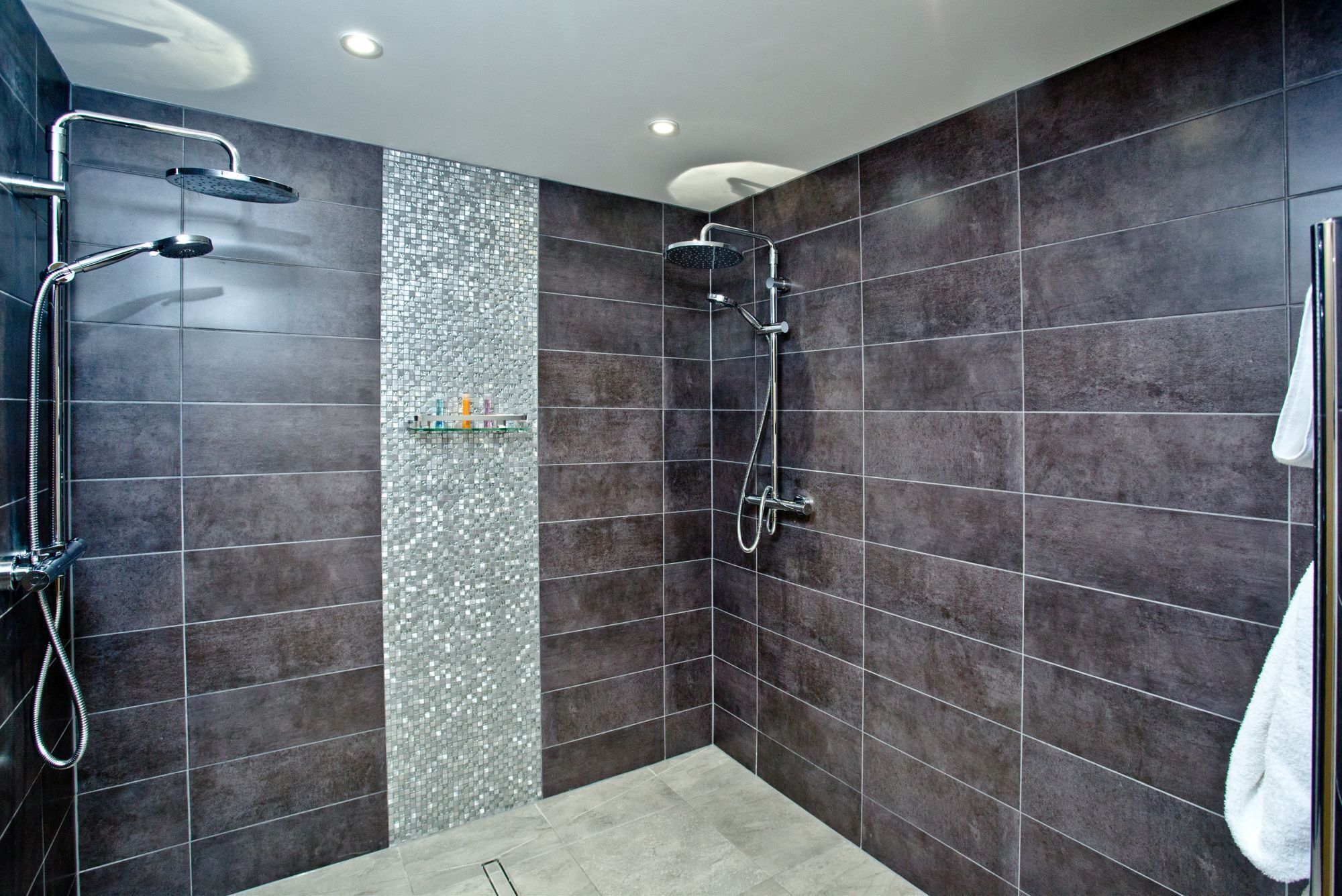 Un box doccia moderno con piastrelle scure, uno scaffale in vetro con articoli da toilette e un asciugamano bianco appeso al muro.