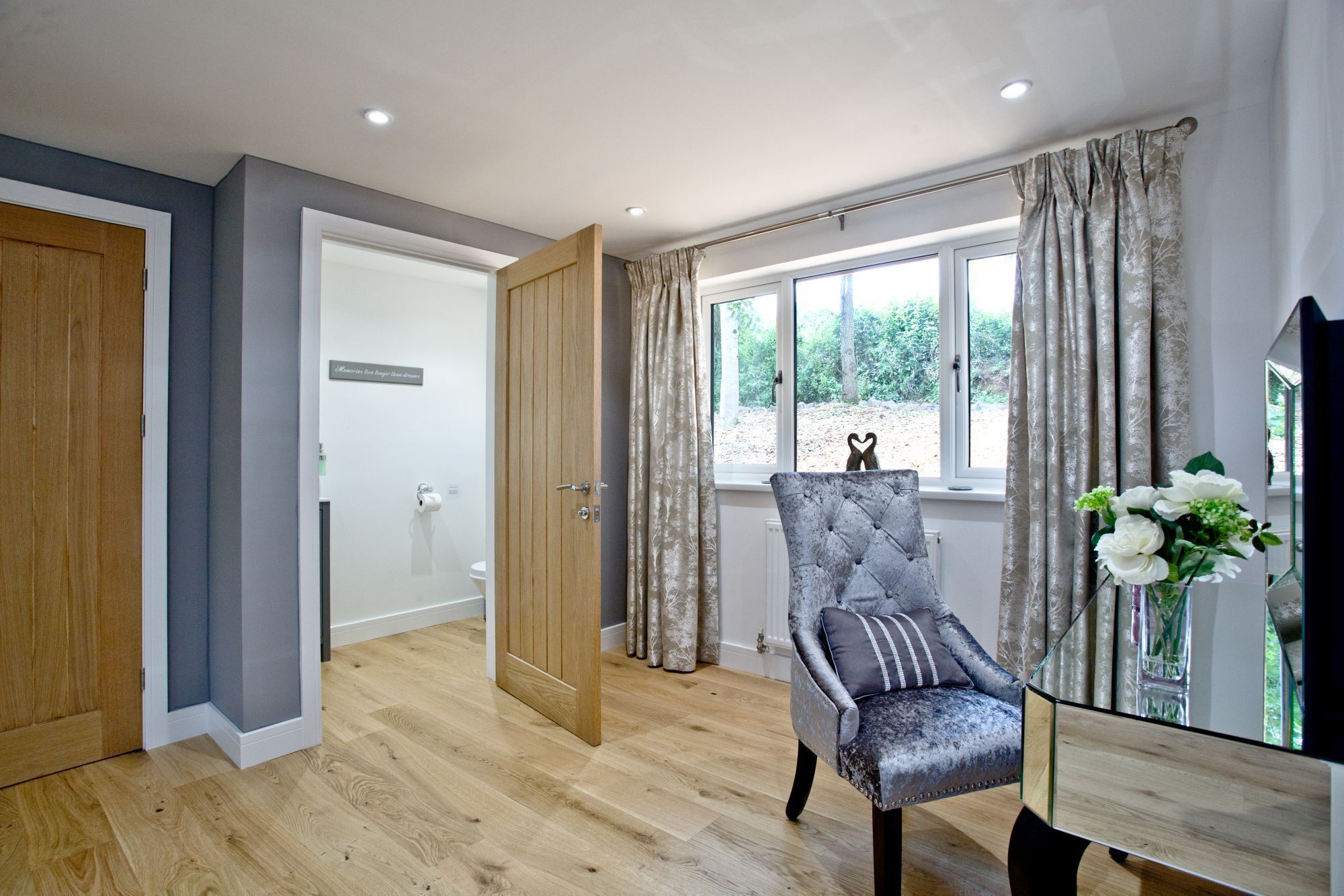 A modern room with a wooden floor, a grey upholstered chair, a glass table, and a window with curtains.