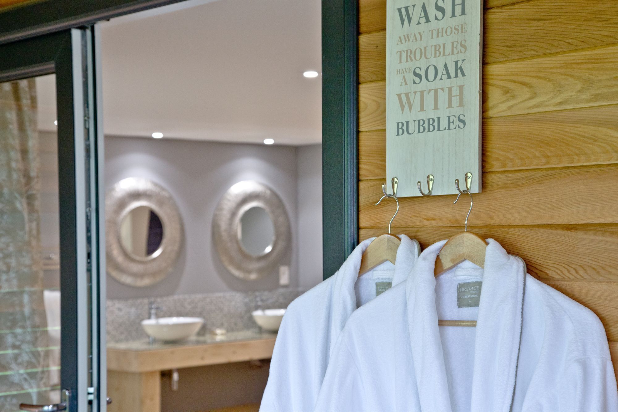 A bathroom with a sign that reads 'WASH AND THOSE TROUBLES HAVE SOAK WITH BUBBLES' and a wooden wall with a towel rack holding white robes.