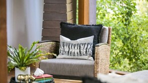 A wicker chair with a grey cushion and a black pillow with a silhouette pattern, placed on a wooden floor with a woven mat and a small potted plant. - Hepburn Spa Pavilions (Hepburn Springs)
