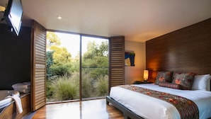 A modern bedroom with a large bed, a built-in TV, and a sliding glass door leading to a balcony with a view of greenery. - Hepburn Spa Pavilions (Hepburn Springs)