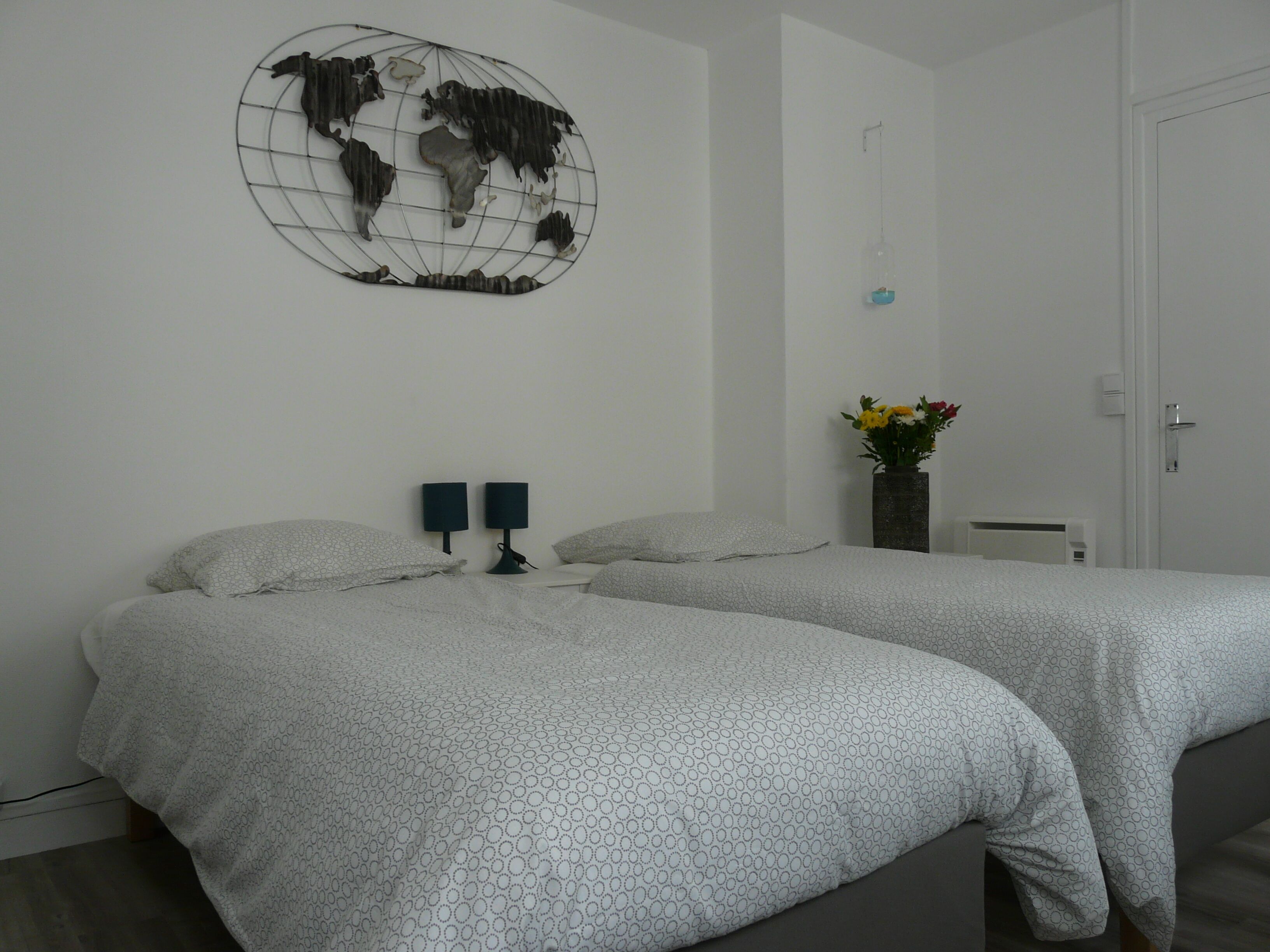 A hotel room with two beds, a globe artwork on the wall, a vase with flowers, and a door.