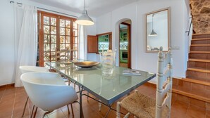 Un comedor con mesa de vidrio, sillas blancas, una lámpara colgante y vista a la cocina.