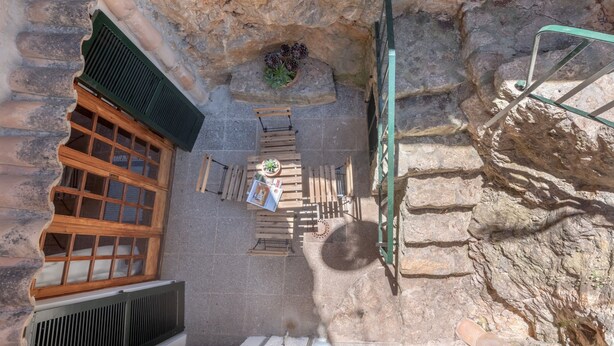 Una habitación con paredes de piedra, una ventana de madera, una escalera metálica verde y una pequeña mesa con una planta.