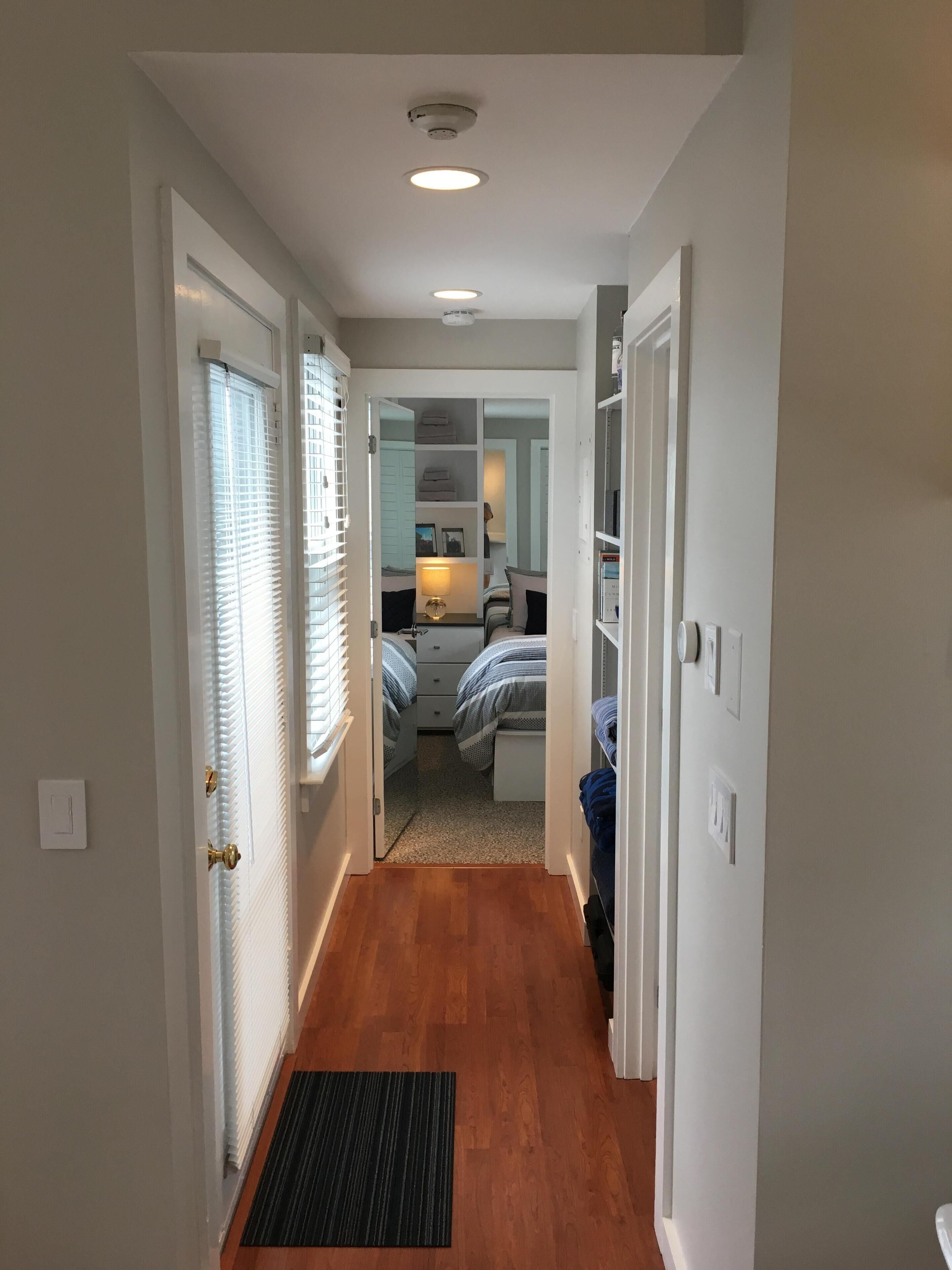 A narrow hallway with a wooden floor leading to a bedroom with a bed and a nightstand.