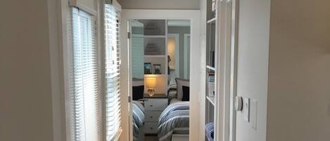 A narrow hallway with a wooden floor leading to a bedroom with a bed and a nightstand.