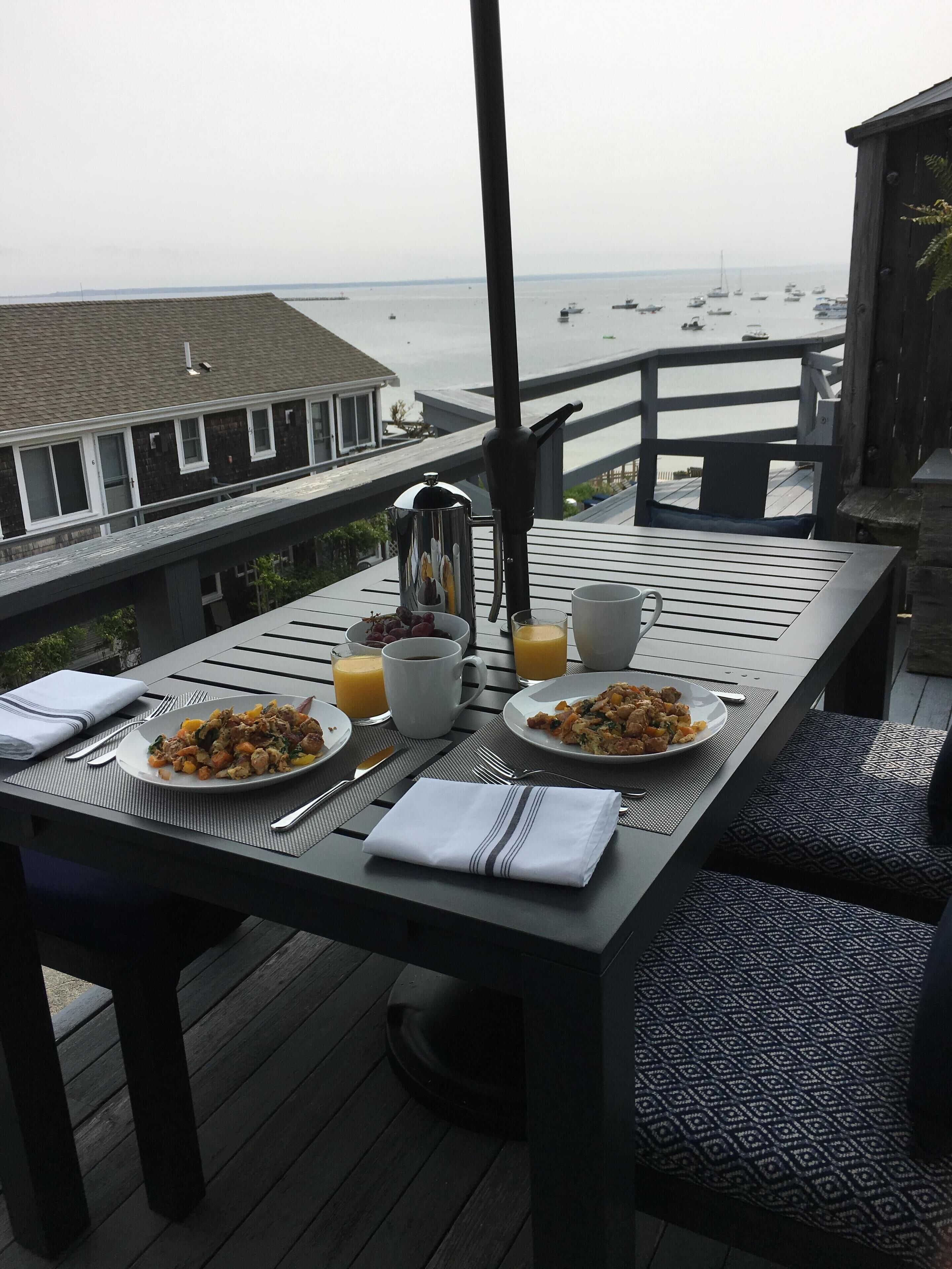 Ein Esstisch mit Frühstücksgeschirr, Tassen und einer Kaffeekanne auf einer Terrasse mit Blick auf eine Marina und Boote.
