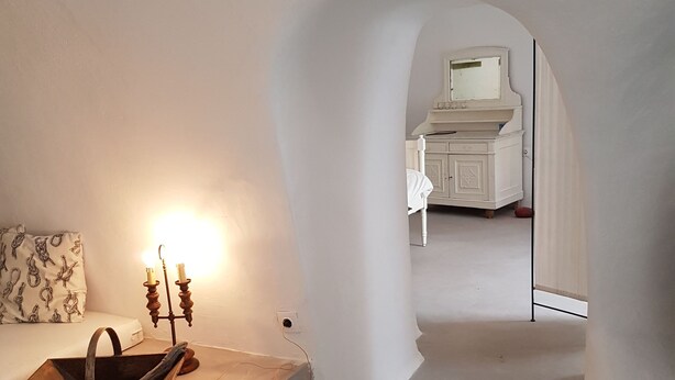 Una habitación con pared blanca, un banco de madera, un candelabro y una cama con cabecero blanco.