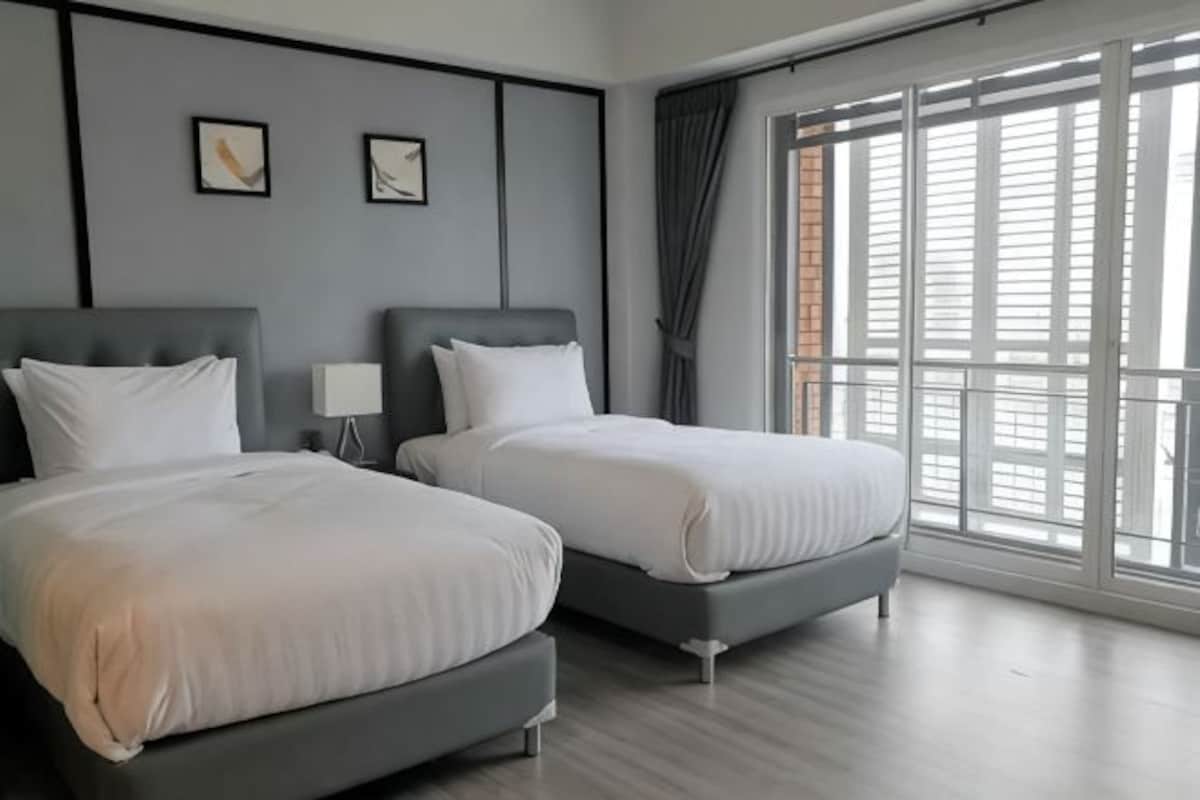 A hotel room with two beds, a nightstand, and a large window with blinds.