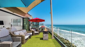 Een balkon met rieten meubilair, een vuurkorf en rode parasols met uitzicht op de oceaan.