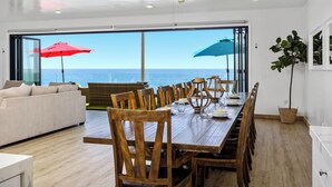 Een eetgedeelte met uitzicht op zee, houten stoelen, een grote tafel en twee parasols.