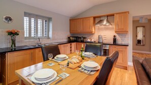A modern kitchen with wooden cabinets, a dining table set for four, and a large window with blinds. - Heron Lodge, Hot Tub, Woodburner (Exeter)