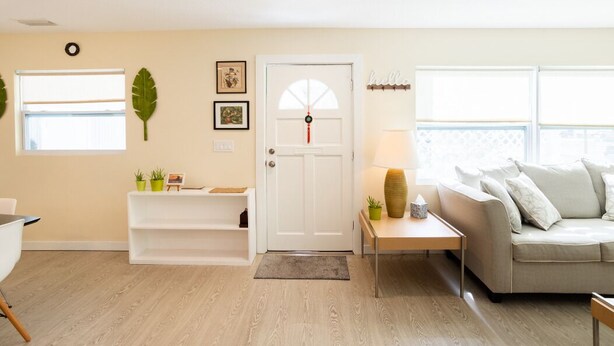 Una sala de estar con una puerta blanca, un sofá, una mesa de centro, una lámpara, un estante pequeño con plantas y una ventana con persianas.