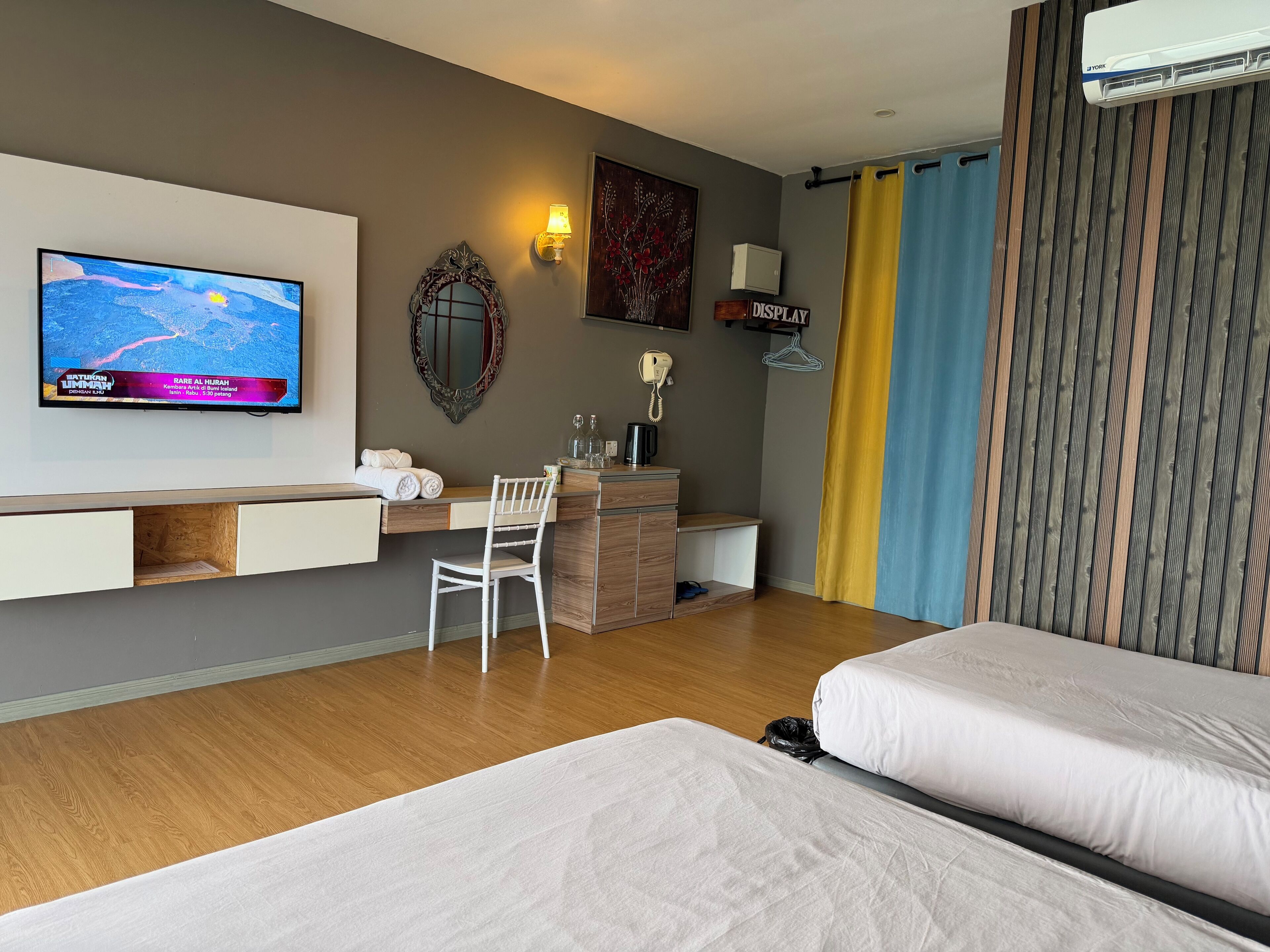 Habitación de hotel con cama, escritorio con silla, televisor mostrando un noticiero y ventana con cortinas.