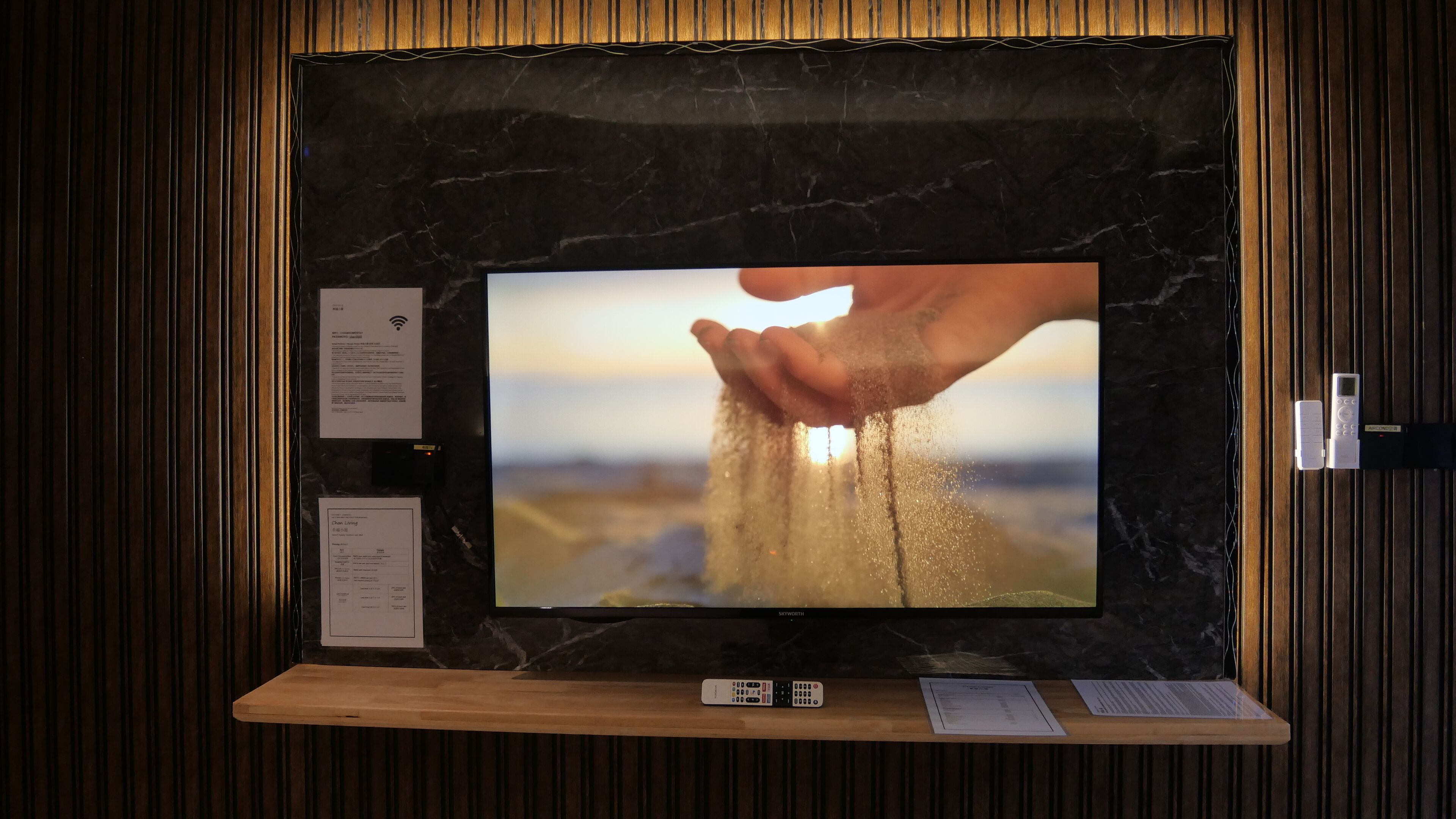 A television mounted on a wall with a wooden panel design, a remote control, and a menu card on the lower shelf.