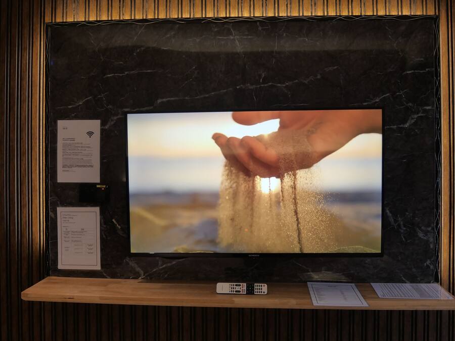 A television mounted on a wall with a wooden panel design, a remote control, and a menu card on the lower shelf.