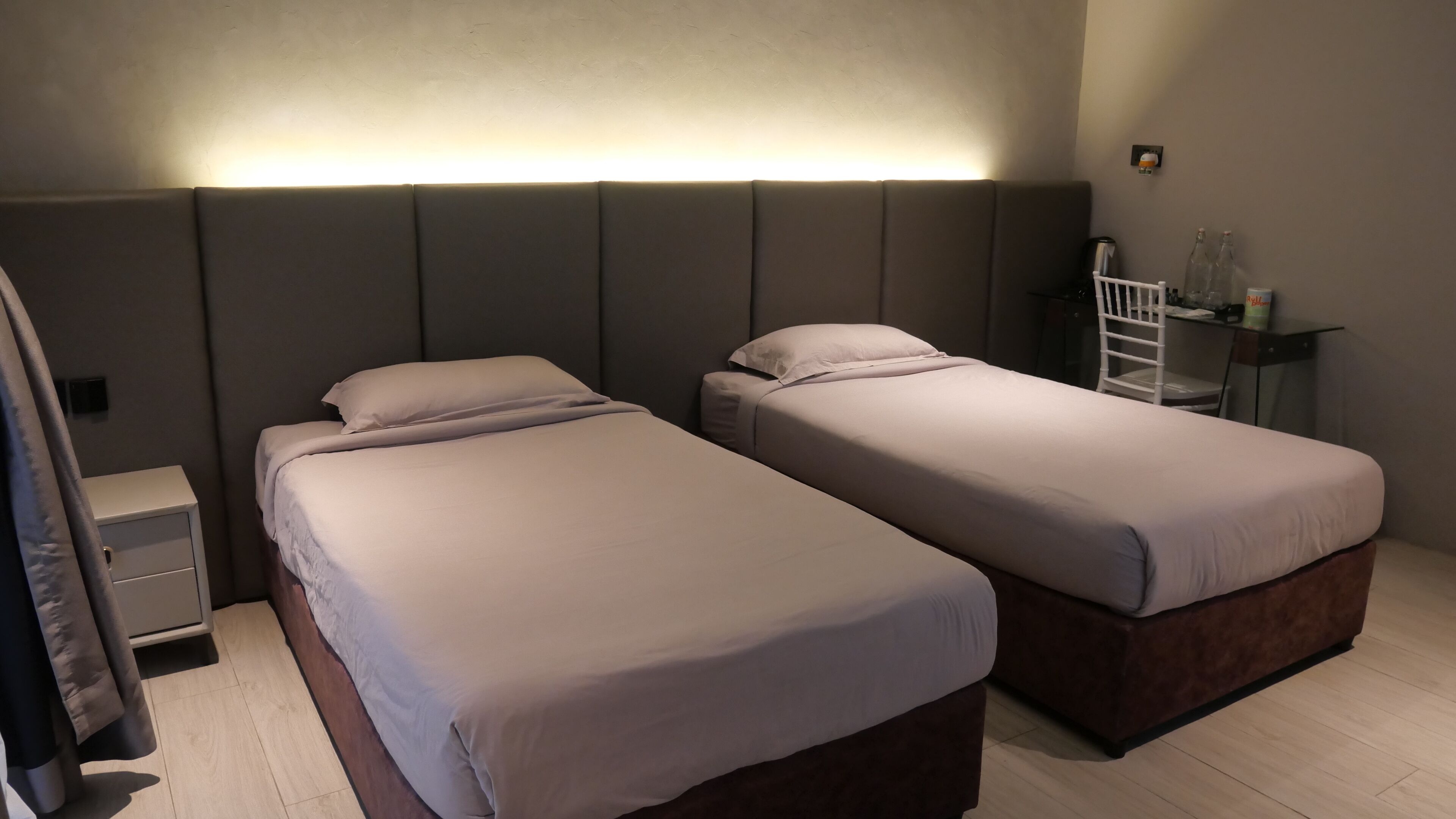 A hotel room with two single beds, a small bedside table, a nightstand, and a wall-mounted shelf with bottles.