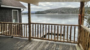 A wooden deck with a railing overlooking a lake, a building with a dark exterior, and a cloudy sky. - Lakefront Retreat on Lake Flower near Lake Placid (Saranac Lake)