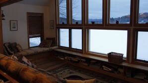 A cozy living room with a large window, a cushioned bench, and a bookshelf. - Lakefront Retreat on Lake Flower near Lake Placid (Saranac Lake)
