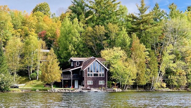 A lakeside cabin with a deck, surrounded by dense forest and a clear sky. - Lakefront Retreat on Lake Flower near Lake Placid (Saranac Lake)
