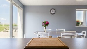 A dining area with a wooden board on the counter, white chairs, a table, a clock on the wall, and a window with a view.