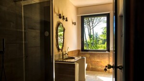 A modern bathroom with a glass shower enclosure, a large mirror, and a window providing a view of greenery.