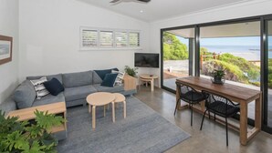 A modern living room with a grey sofa, wooden coffee table, and a dining area with a wooden table and chairs. There is a TV on a stand and a large window with a view of the sea. - FortyTwo Mini |Gracetown (Gracetown)
