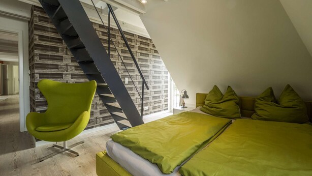 Un dormitorio con techo inclinado, una silla verde, una cama con ropa de cama verde y una pared con diseño de ladrillo.