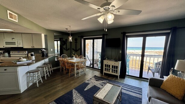 Un espacio habitable amplio con una mesa de comedor, un ventilador de techo y vistas a la playa a través de grandes puertas correderas de vidrio.