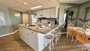 Una cocina con isla central, armarios blancos, microondas y un comedor con sillas y mesa de madera.