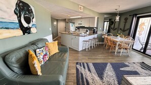 Una sala moderna con un sofá verde, una zona de comedor con sillas de madera, una cocina con armarios blancos y un cuadro grande de un perro en la pared.