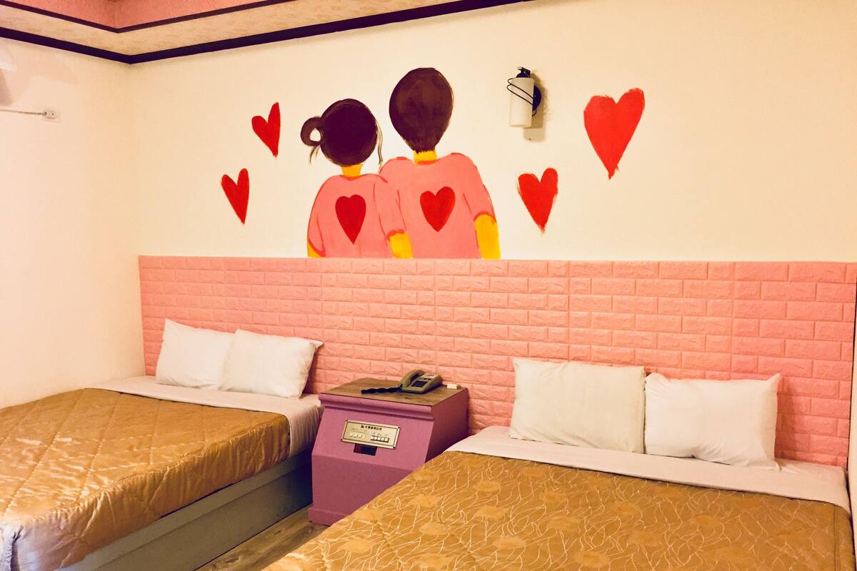 A hotel room with two beds, each with a pink headboard and white bedding. There is a wall decoration of two figures with hearts above them.