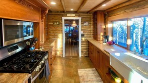 A log cabin kitchen with a stainless steel stove, wooden cabinets, and a large window overlooking a forest.