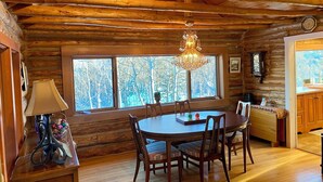 A rustic log cabin interior with a dining table, chairs, a chandelier, a large window, and a clock on the wall.