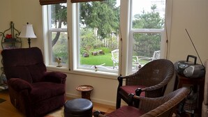 A living room with a large window, a red armchair, a wicker chair, a small round table, and a radio on a wooden side table.