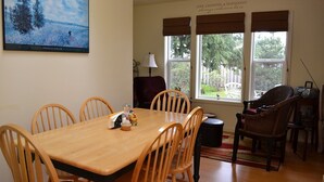 A dining room with a wooden table and chairs, a painting on the wall, and a view of the outdoors through large windows.