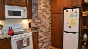 A kitchen with a white microwave, stove, and refrigerator, wooden cabinets, and a brick wall.