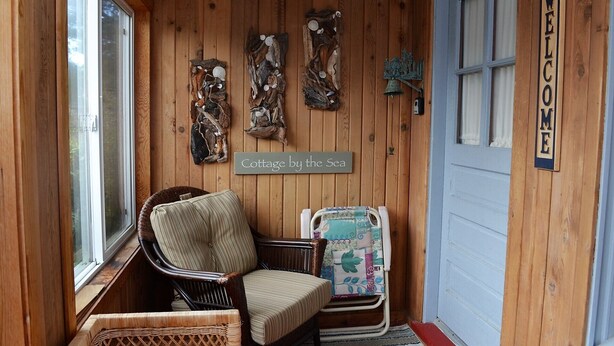 A wooden cabin with a wicker chair, a sign reading "Cottage by the Sea", and a "WELCOME" sign on the door.